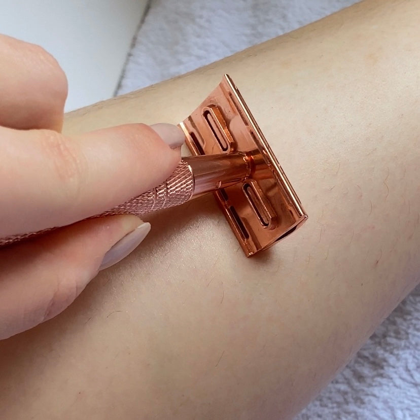 Fotografia prezintă o mână feminină cu unghii îngrijite, aplicând un aparat de ras manual cu două tăișuri pe pielea brațului. Aparatul de ras are un finisaj metalic roz-auriu, și este ținut cu mânerul texturat în relief pentru o aderență sigură. Detaliul și calitatea aparatului de ras sunt evidențiate prin contrastul cu pielea și fundalul textil alb. Imaginea captează un moment intim de îngrijire personală, cu o focalizare clară pe aparatul de